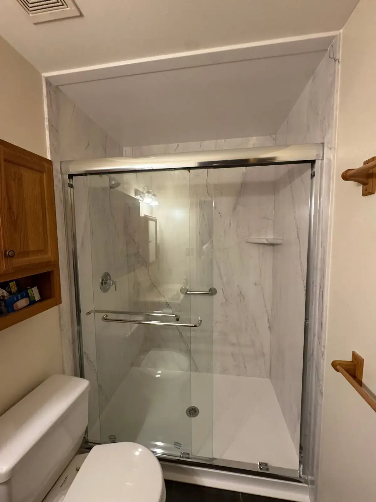 A finished walk-in shower with white marble-look walls and a chrome sliding glass door in a small bathroom with wood cabinets